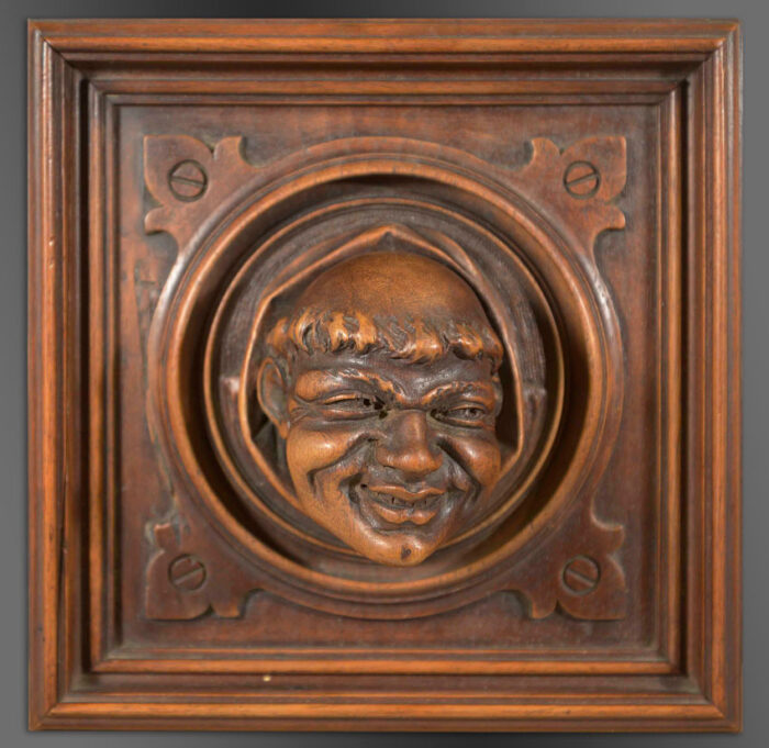 Trompe L'oeil carved in wood depicting a monk's head peeping out of a circular architectural roundel set within a square panel.